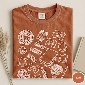 Puede incluir: Una camiseta de color &oacute;xido con un gr&aacute;fico de l&iacute;neas blancas de varias formas de pasta, incluyendo farfalle, raviolis, penne y l&aacute;minas de lasa&ntilde;a. La camiseta tiene un cuello redondo y una etiqueta de la marca Comfort Colors en el interior.