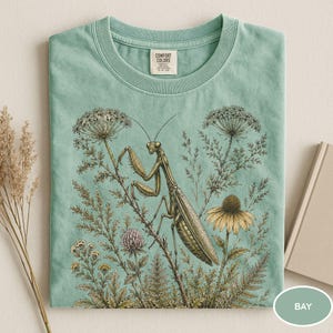 May include: A light green t-shirt featuring a detailed illustration of a praying mantis amidst wildflowers and foliage. The design includes a yellow coneflower, a pink clover, and various botanical elements. The shirt has a crew neck and a tag that says "Comfort Colors".
