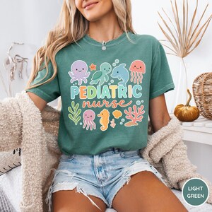 K&ouml;nnte beinhalten: Hellgr&uuml;nes T-Shirt mit der Aufschrift "PEDIATRIC nurse" in bunten Buchstaben und Meereskreaturen-Grafiken. Das Design umfasst einen Oktopus, eine Schildkr&ouml;te, einen Delfin, eine Qualle, ein Seepferdchen und Korallen. Das Shirt ist aus weichem Material.