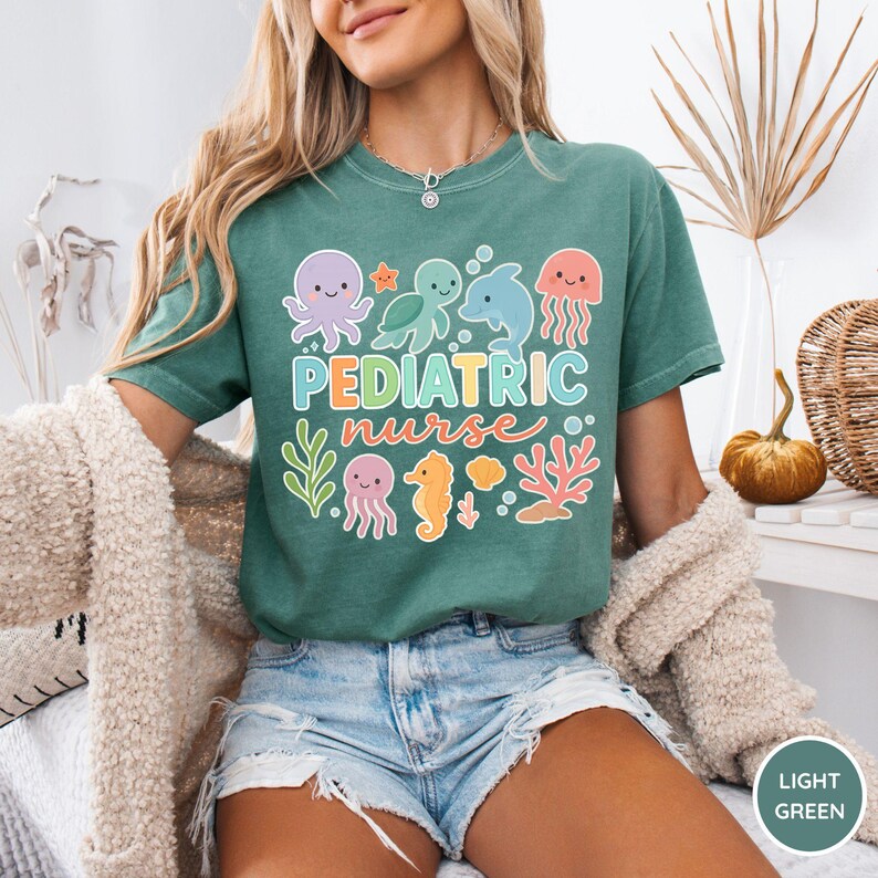 K&ouml;nnte beinhalten: Hellgr&uuml;nes T-Shirt mit der Aufschrift "PEDIATRIC nurse" in bunten Buchstaben und Meereskreaturen-Grafiken. Das Design umfasst einen Oktopus, eine Schildkr&ouml;te, einen Delfin, eine Qualle, ein Seepferdchen und Korallen. Das Shirt ist aus weichem Material.