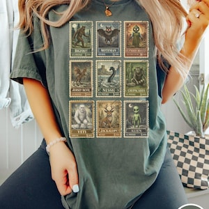 May include: Dark green t-shirt featuring a collection of vintage-style postage stamps with cryptid illustrations. The stamps depict creatures like Bigfoot, Mothman, and the Jersey Devil. The shirt has a relaxed fit and the word "MOSS" is printed on a circle.