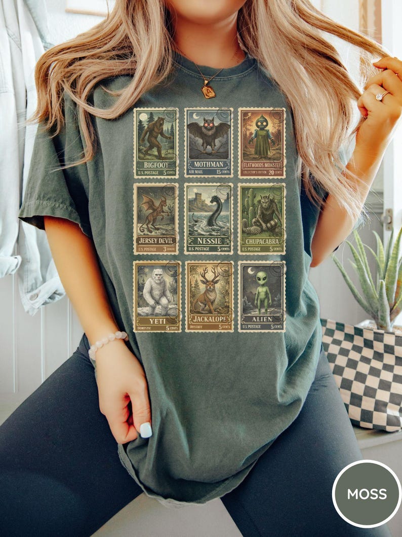 May include: Dark green t-shirt featuring a collection of vintage-style postage stamps with cryptid illustrations. The stamps depict creatures like Bigfoot, Mothman, and the Jersey Devil. The shirt has a relaxed fit and the word "MOSS" is printed on a circle.