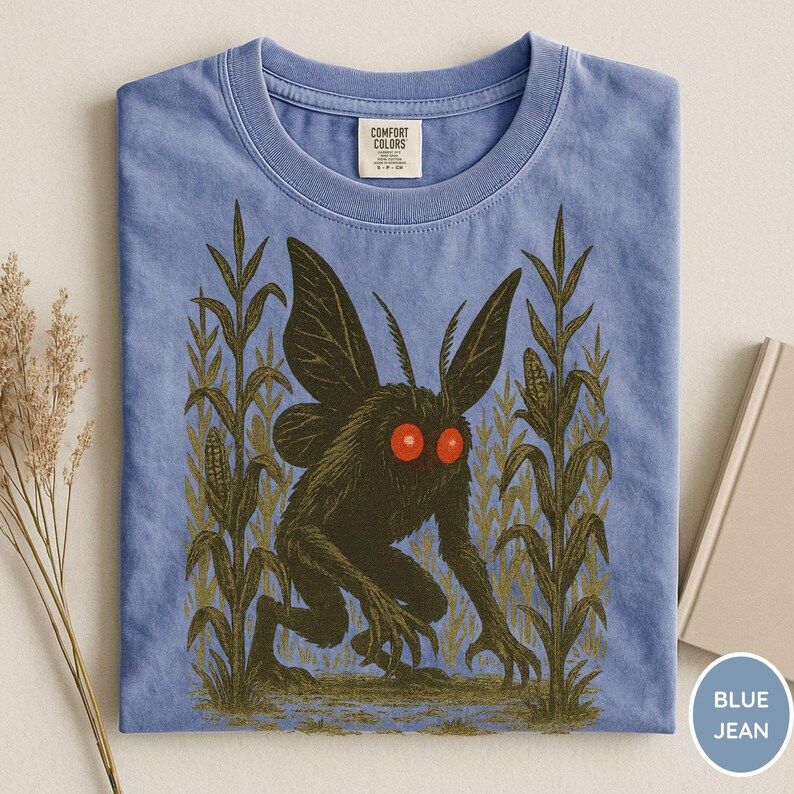 K&ouml;nnte beinhalten: Blaues Jeans-T-Shirt mit einer dunklen Kreatur mit roten Augen und gro&szlig;en Fl&uuml;geln, die in einem Maisfeld steht. Das Grafikdesign ist auf ein T-Shirt der Marke Comfort Colors gedruckt. Das Shirt hat einen klassischen Rundhalsausschnitt.