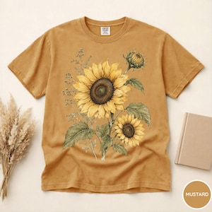 May include: Mustard-colored t-shirt featuring a detailed watercolor illustration of sunflowers. The design includes two large sunflowers, buds, and foliage. The shirt has a classic crew neck and short sleeves. The tag reads "COMFORT COLORS".