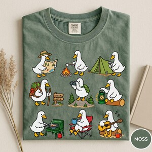 May include: A moss green t-shirt featuring a graphic print of nine white ducks engaged in various camping activities, including reading a map, roasting a marshmallow, setting up a tent, hiking, using binoculars, holding a lantern, cooking, sitting in a chair, and playing guitar.