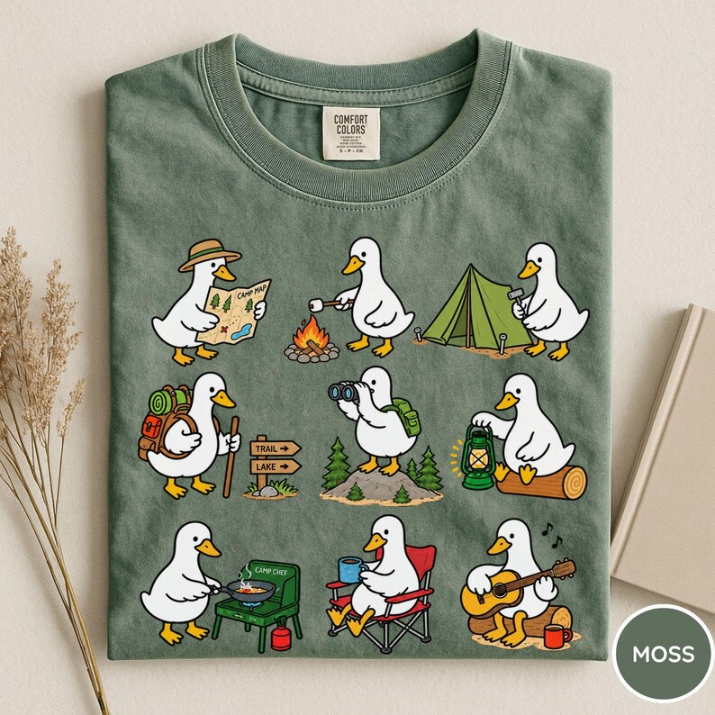 May include: A moss green t-shirt featuring a graphic print of nine white ducks engaged in various camping activities, including reading a map, roasting a marshmallow, setting up a tent, hiking, using binoculars, holding a lantern, cooking, sitting in a chair, and playing guitar.
