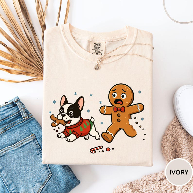 May include: Ivory-colored sweatshirt featuring a cartoon French Bulldog in a Christmas sweater, holding a gingerbread man's leg, and a frightened gingerbread man. Snowflakes and a candy cane are also depicted. The shirt is folded.