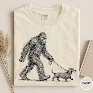 May include: Ivory-colored t-shirt featuring a black and white illustration of Bigfoot walking a dachshund on a leash. The graphic is a detailed pen and ink style drawing. The shirt has a crew neck and short sleeves.