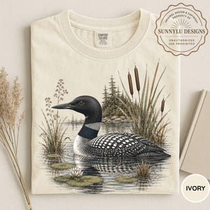 May include: An ivory t-shirt featuring a detailed illustration of a black and white loon swimming in a lake. The graphic includes cattails, pine trees, and water lilies. The shirt has a Comfort Colors label at the neck and a SunnyLu Designs logo.