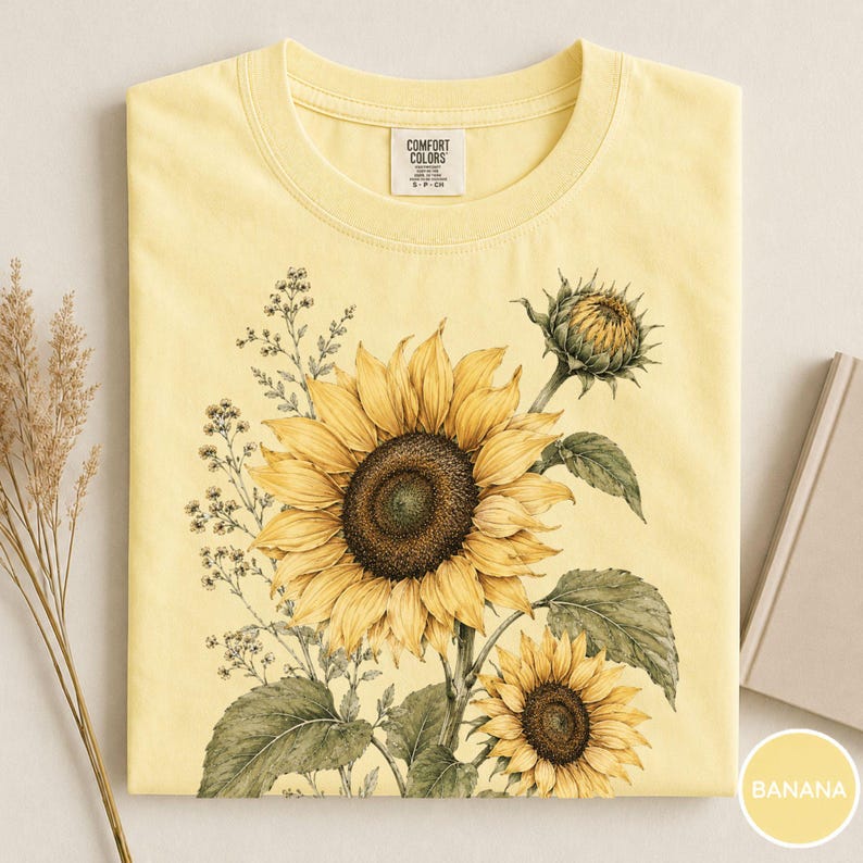 May include: A yellow Comfort Colors t-shirt featuring a detailed illustration of sunflowers in various stages of bloom. The design includes a large, open sunflower, a smaller one, and a bud, with green leaves and delicate sprigs. The shirt is folded neatly.