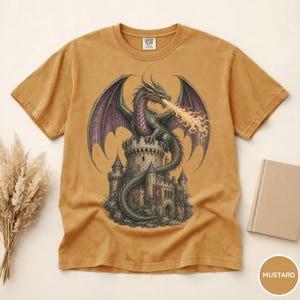 May include: A mustard yellow short-sleeve t-shirt featuring a graphic print of a green dragon perched atop a stone castle tower. The dragon has large purple wings and is breathing fire. The shirt has a crew neckline and a Comfort Colors brand label.