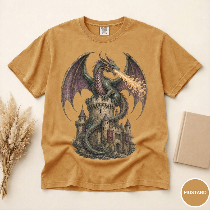 May include: A mustard yellow short-sleeve t-shirt featuring a graphic print of a green dragon perched atop a stone castle tower. The dragon has large purple wings and is breathing fire. The shirt has a crew neckline and a Comfort Colors brand label.