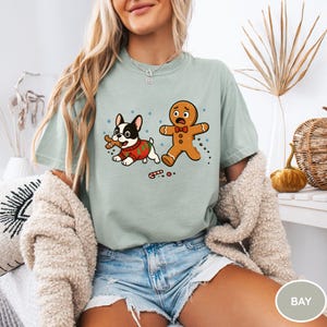 May include: A light green t-shirt featuring a cartoon illustration of a French Bulldog in a Christmas sweater holding a bone, and a scared gingerbread man. The design includes snowflakes and candy canes.