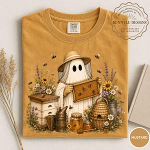 May include: A mustard yellow t-shirt featuring a graphic of a ghost wearing a beekeeper hat and veil, holding a honeycomb frame. The design includes a beehive, a smoker, a honey jar, and various flowers with bees. The label reads Comfort Colors.