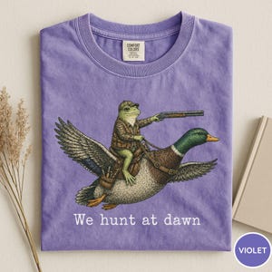Puede incluir: Camiseta violeta Comfort Colors con una ilustraci&oacute;n humor&iacute;stica de una rana con atuendo de caza montada en un pato y sosteniendo una escopeta. El texto "We hunt at dawn" est&aacute; impreso debajo. La camiseta tiene cuello redondo.