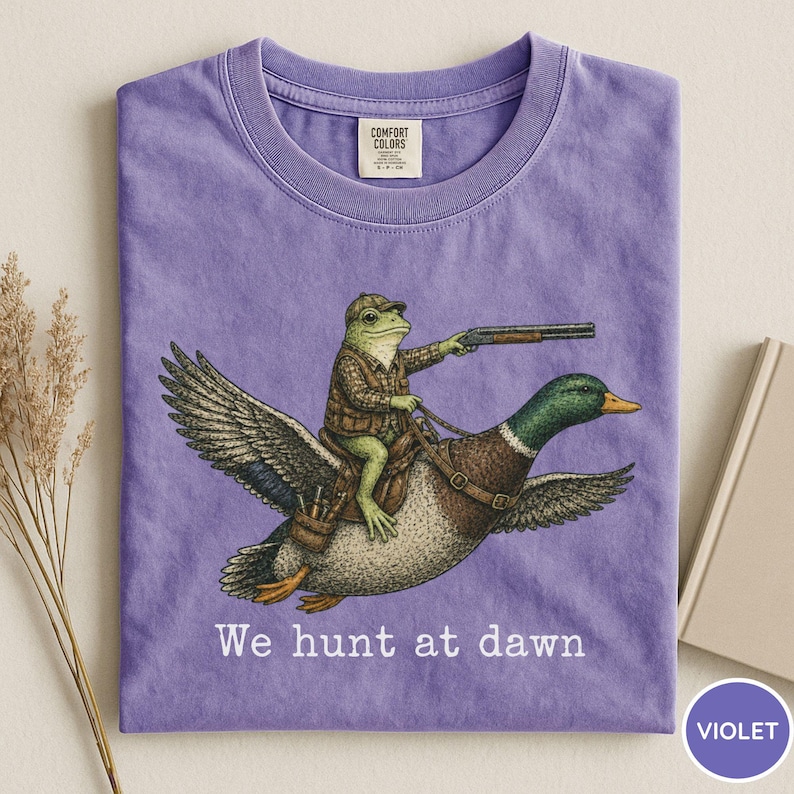 Puede incluir: Camiseta violeta Comfort Colors con una ilustraci&oacute;n humor&iacute;stica de una rana con atuendo de caza montada en un pato y sosteniendo una escopeta. El texto "We hunt at dawn" est&aacute; impreso debajo. La camiseta tiene cuello redondo.