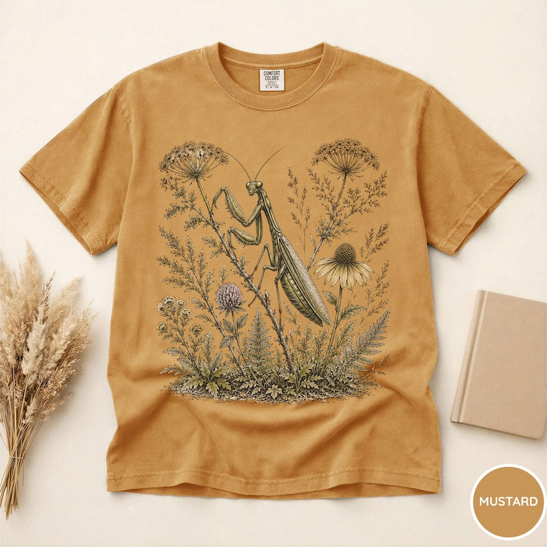 May include: Mustard-colored t-shirt featuring a detailed illustration of a praying mantis perched among wildflowers and foliage. The design includes various plants and a detailed insect, creating a nature-inspired aesthetic. The shirt has a classic crew neck and short sleeves.