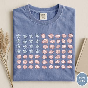 May include: A blue short-sleeve t-shirt with a crew neckline and a Comfort Colors label. The front features a graphic design of white starfish and red seashells arranged in a pattern resembling the American flag. The fabric has a washed texture.