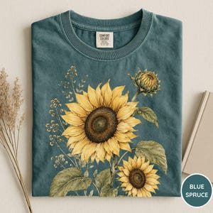 May include: A blue spruce t-shirt featuring a watercolor illustration of sunflowers. The design includes a large, fully bloomed sunflower, a smaller one, and a sunflower bud, with green leaves and stems. The shirt has a crew neck and a tag that says "Comfort Colors".