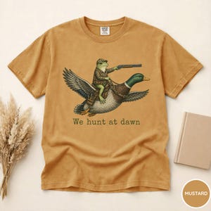 Puede incluir: Camiseta de color mostaza con una ilustraci&oacute;n caprichosa. Una rana con atuendo de caza monta un pato, sosteniendo un rifle. El texto "We hunt at dawn" est&aacute; impreso debajo. La camiseta tiene cuello redondo cl&aacute;sico y mangas cortas.