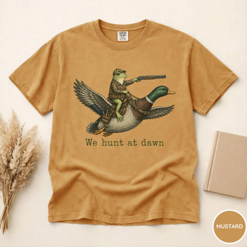 Puede incluir: Camiseta de color mostaza con una ilustraci&oacute;n caprichosa. Una rana con atuendo de caza monta un pato, sosteniendo un rifle. El texto "We hunt at dawn" est&aacute; impreso debajo. La camiseta tiene cuello redondo cl&aacute;sico y mangas cortas.