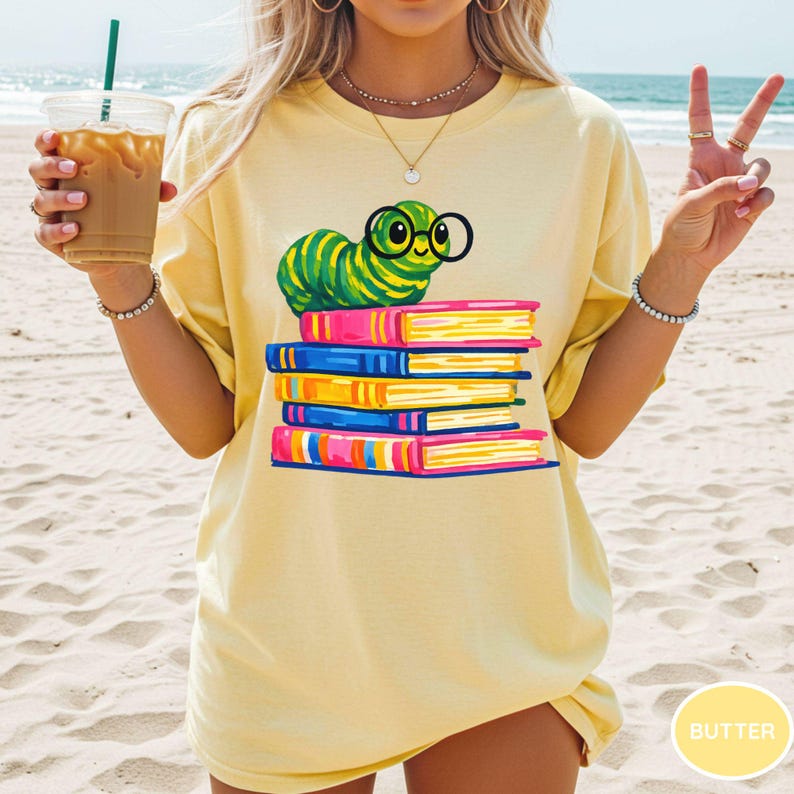Pu&ograve; includere: Una t-shirt giallo pallido con la grafica di un bruco verde che indossa occhiali tondi neri, appoggiato sopra una pila di cinque libri colorati nelle tonalit&agrave; del rosa, blu e giallo. La t-shirt ha un girocollo e maniche corte.