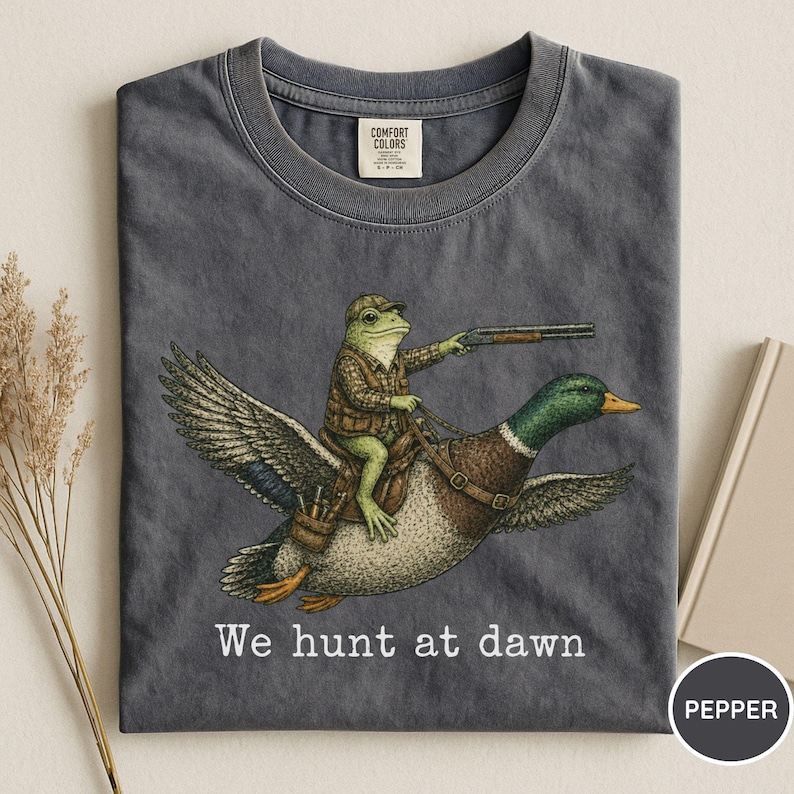 Puede incluir: Camiseta gris oscuro con una ilustraci&oacute;n caprichosa de una rana con atuendo de caza montada en un pato y sosteniendo una escopeta. El texto "We hunt at dawn" est&aacute; impreso debajo. Marca Comfort Colors.