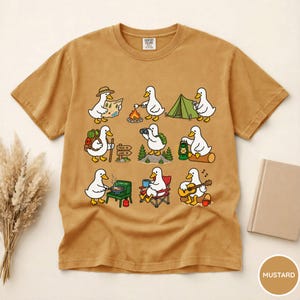 May include: A mustard yellow short-sleeve t-shirt featuring a graphic print of nine white ducks engaged in various camping activities, including hiking, roasting marshmallows, setting up a tent, cooking, and playing an acoustic guitar.