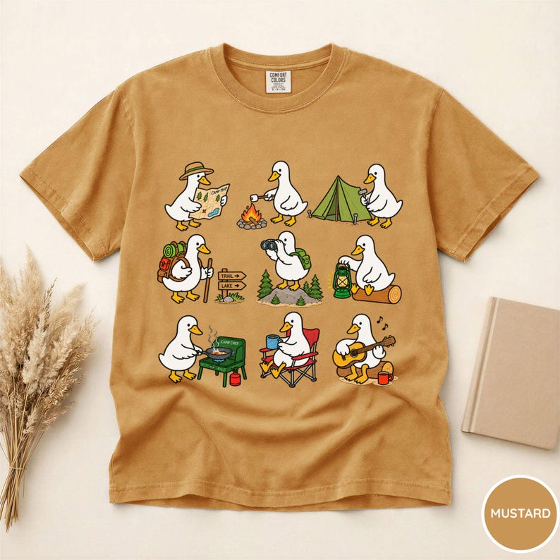 May include: A mustard yellow short-sleeve t-shirt featuring a graphic print of nine white ducks engaged in various camping activities, including hiking, roasting marshmallows, setting up a tent, cooking, and playing an acoustic guitar.