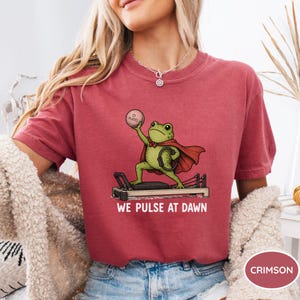 May include: A crimson t-shirt featuring a graphic of a green frog wearing a red cape, standing on a pilates reformer machine while holding a pink pilates ball. The text below the graphic reads WE PULSE AT DAWN in white capital letters.