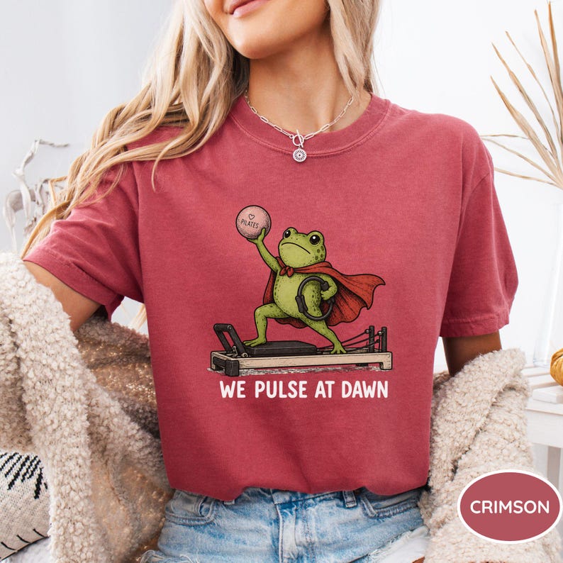 May include: A crimson t-shirt featuring a graphic of a green frog wearing a red cape, standing on a pilates reformer machine while holding a pink pilates ball. The text below the graphic reads WE PULSE AT DAWN in white capital letters.