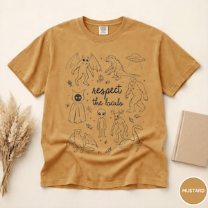 May include: Mustard-colored t-shirt with a black graphic featuring various cryptids and the text "respect the locals." The design includes a winged creature, a dinosaur, a UFO, a Bigfoot, an alien, and the Loch Ness Monster.