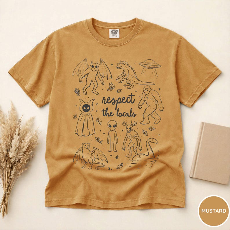 May include: Mustard-colored t-shirt with a black graphic featuring various cryptids and the text "respect the locals." The design includes a winged creature, a dinosaur, a UFO, a Bigfoot, an alien, and the Loch Ness Monster.