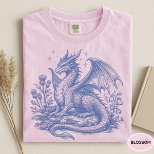 Peut inclure: T-shirt rose clair avec un dessin bleu d'un dragon ail&eacute;, entour&eacute; de fleurs et d'&eacute;toiles. Le t-shirt est &eacute;tiquet&eacute; "Blossom" et porte une &eacute;tiquette Comfort Colors.