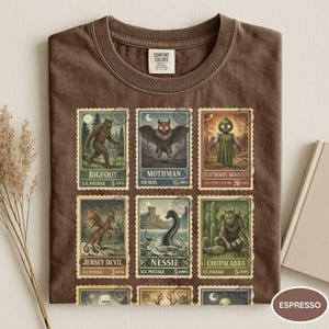 May include: Espresso-colored t-shirt featuring a collection of vintage-style postage stamps. The stamps depict various cryptids, including Bigfoot, Mothman, and the Flatwoods Monster. The shirt is made of a soft material.