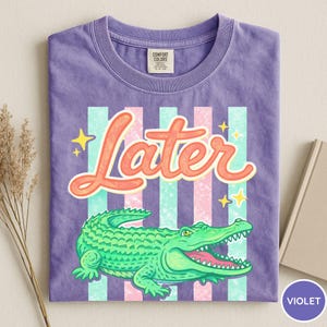May include: Violet t-shirt with a graphic of a green alligator and the word "Later" in orange script. The design includes pink and teal stripes and yellow stars. The shirt is folded.
