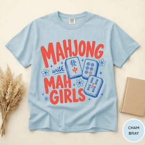 May include: A light blue short-sleeve t-shirt featuring red text that reads MAHJONG with MAH GIRLS and the word with in blue. The design includes three blue and white mahjong tiles, small blue flower icons, and star accents on the front.