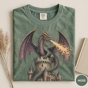 May include: A sage green t-shirt featuring a graphic print of a dragon perched atop a stone castle tower, breathing a stream of yellow fire. The dragon has purple wings and textured scales. The shirt has a Comfort Colors brand label at the neck.