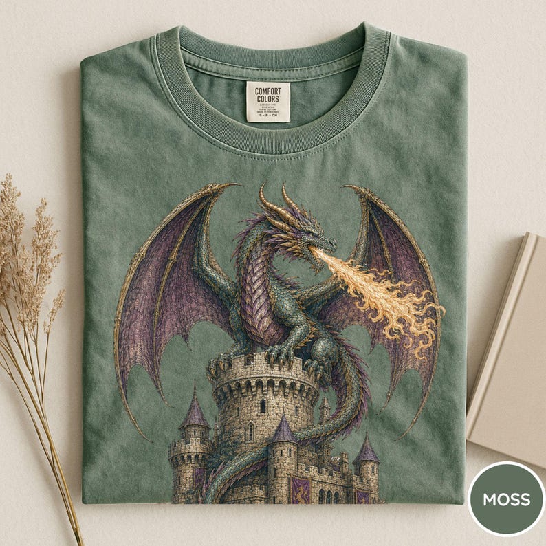 May include: A sage green t-shirt featuring a graphic print of a dragon perched atop a stone castle tower, breathing a stream of yellow fire. The dragon has purple wings and textured scales. The shirt has a Comfort Colors brand label at the neck.