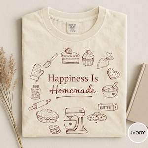 May include: Ivory-colored t-shirt with a crew neck. The shirt features a brown graphic of baking-related items, including a rolling pin, mixer, and pie, surrounding the text "Happiness Is Homemade."