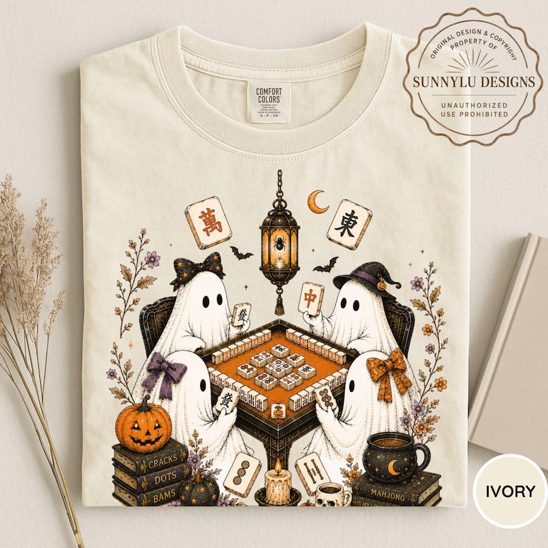 May include: An ivory t-shirt featuring a graphic of four ghosts playing mahjong around a table. The design includes a hanging lantern, a pumpkin, stacks of books labeled Cracks, Dots, and Bams, and various mahjong tiles with Chinese characters.
