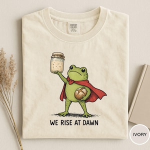 May include: Ivory-colored t-shirt featuring a cartoon frog superhero holding a jar and a loaf of bread. The frog wears a red cape and a bow tie. The text "WE RISE AT DAWN" is printed below the frog.