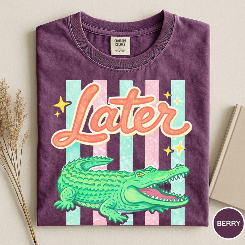 May include: Berry-colored t-shirt with a graphic of a green alligator and the word "Later" in orange script. The design includes vertical stripes in light blue and pink, with yellow star accents. The shirt is folded.