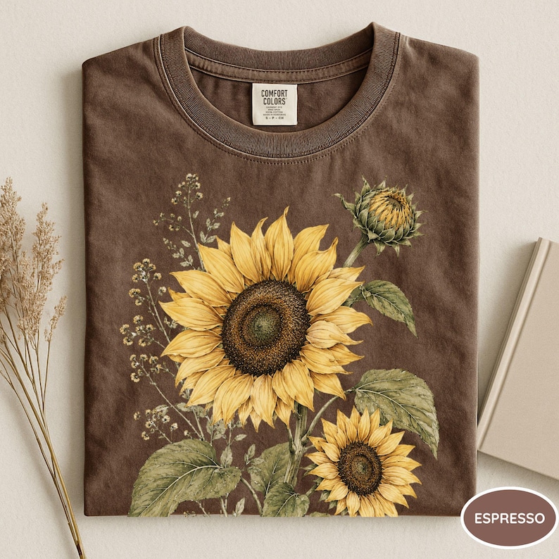May include: Espresso-colored t-shirt featuring a watercolor sunflower design. The graphic includes a large, open sunflower with yellow petals and a dark brown center, along with smaller sunflowers and green leaves. The shirt has a crew neck and a tag that says "Comfort Colors."