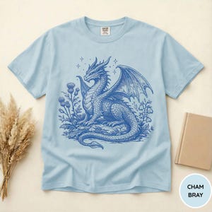 Peut inclure: T-shirt bleu clair avec une illustration d&eacute;taill&eacute;e d'un dragon bleu, avec des ailes, des fleurs et des &eacute;toiles. Le t-shirt a un col rond et des manches courtes. Le texte "CHAM BRAY" est dans un cercle en bas &agrave; droite.