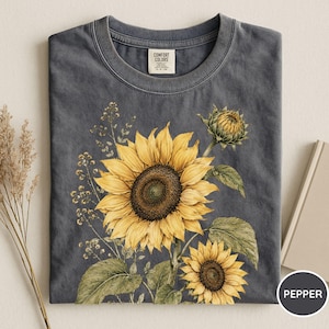 May include: A dark gray t-shirt featuring a watercolor illustration of sunflowers. The design includes a large, fully bloomed sunflower, a smaller one, and a sunflower bud, with green leaves and stems. The shirt has a crew neck.