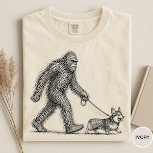 May include: Ivory-colored t-shirt featuring a black and white illustration of Bigfoot walking a corgi on a leash. The shirt has a crew neck and the Comfort Colors brand label is visible. The word "IVORY" is printed on the bottom right.