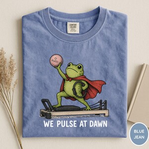 May include: A blue t-shirt featuring a green frog wearing a red cape, standing on a pilates reformer machine. The frog holds a pink ball labeled "PILATES" and a black ring. Below the graphic, the text reads "WE PULSE AT DAWN" in white.