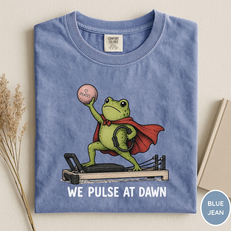 May include: A blue t-shirt featuring a green frog wearing a red cape, standing on a pilates reformer machine. The frog holds a pink ball labeled "PILATES" and a black ring. Below the graphic, the text reads "WE PULSE AT DAWN" in white.