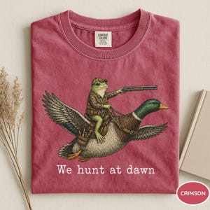 Puede incluir: Camiseta de color carmes&iacute; con una ilustraci&oacute;n humor&iacute;stica de una rana con ropa de caza montada en un pato y sosteniendo una escopeta. El texto "We hunt at dawn" est&aacute; impreso debajo. La camiseta es de material suave.